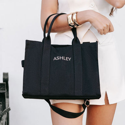 Person holding a black handbag with 'ASHLEY' branding in a room with a fireplace.